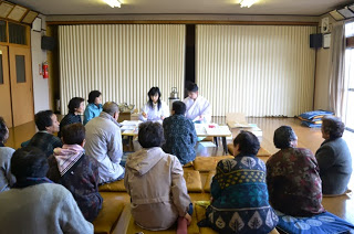 室内に地域住民の方々集まって座っており、前方に置かれた長机に白衣と袴姿の神職と関係者のに女性が座っている室内の様子を写した写真