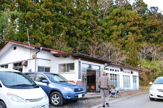 杉林を背景に、集会所のような建物の前に車が数台停まっており、住民が建物に向かって歩いている様子の写真
