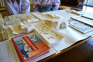 神職と関係者の女性が座る机の上に並べられた真新しいお札や御神像、紙垂(しで)、お供物をかたどった「きりこ」などが並べられている様子の意写真