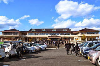 駐車場に多くの車が停まり、えんじ色の屋根でベージュ色の外壁の校舎に向かって生徒や保護者が歩いている様子の写真