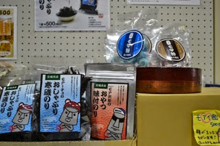 モアイのイラストが描かれた青いパッケージの「おじゃぶり養殖のり」、隣に赤いパッケージの「おやつ味付けのり」、その隣に箱に置かれた「モイアイ飴 500円」が陳列されている写真