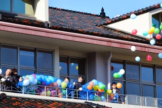 大きな窓と瓦屋根の校舎のバルコニーから、色とりどりの風船が空に放たれている様子の写真