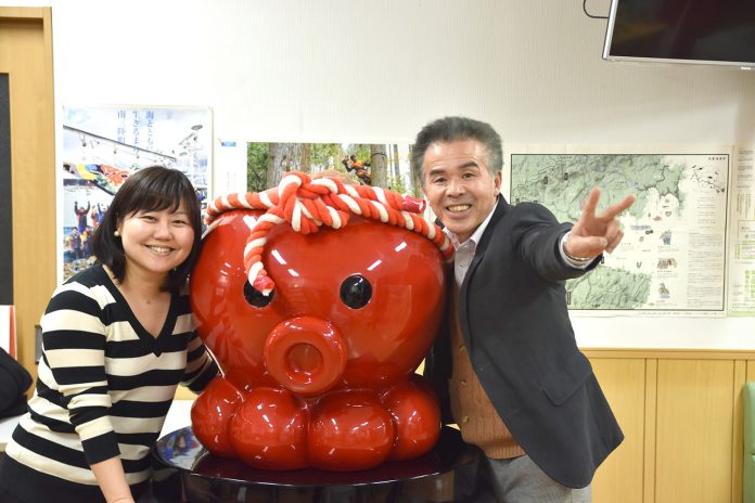 紅白のねじり鉢巻きを巻いた赤いタコのキャラクター「オクトパス君」の像をはさんで、女性リポーターと、ピースサインをした阿部さんが立っている写真