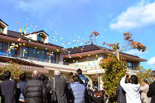 紅葉に彩られた木々に囲まれた校舎の前で、色とりどりの風船が空に放たれている様子を大勢の卒業生や在校生が見上げている写真