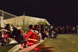 道路脇に座った多くの観客が、夜空を見上げながら花火を鑑賞している様子を側面から捉えた写真