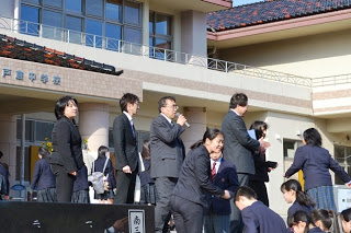 戸田中学校の校舎前で、スーツ姿の卒業生や制服姿の在校生が校舎に向かって歩いているなか、中央に立つ男性が右手でマイクを持ち話をしている様子の写真