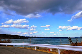 手前から雨雲が現れた空の下、白いガードレールに沿って海沿いを走る道路が、青空と遠くの山々に向かって続く風景の写真