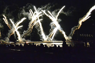 水辺の前に座った観客たちのシルエット越しに、大きな打ち上げ花火が夜空にX字型に広がっている写真