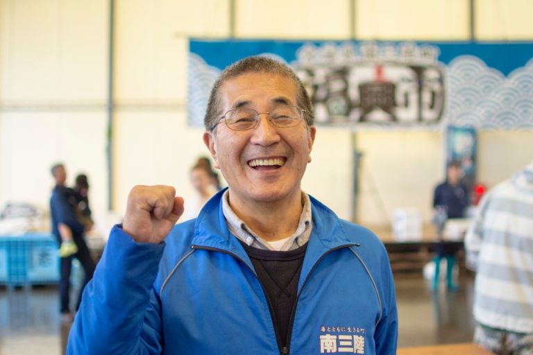 左胸に白色で南三陸と書かれた青色のジャケットを着た山内さんが、笑顔でガッツポーズをしている様子をアップで撮影した写真