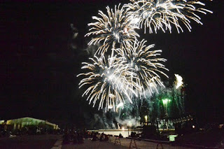 水辺の上空に白い大きな花火がいくつも重なって打ち上がり、夜空が一気に明るくなる瞬間をとらえた写真