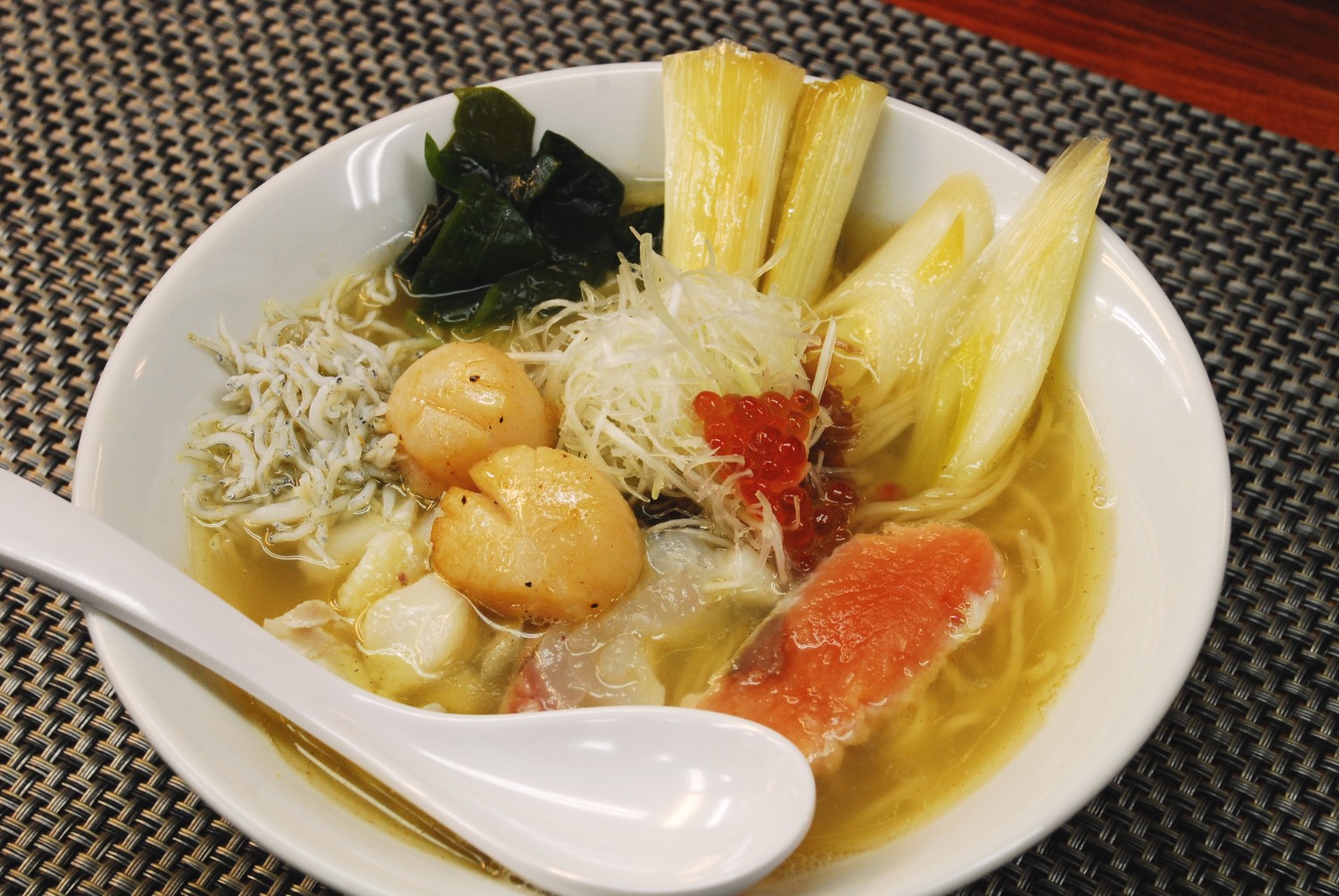 白いラーメン鉢に、ホタテ、いくら、わかめ、ネギ、しらすなど海の幸と野菜がたっぷりのったラーメンが盛り付けられている写真