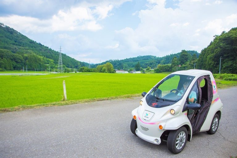 緑が広がる田んぼと山々に囲まれた田舎道を、一人乗りの小型電気自動車が走っている風景の写真