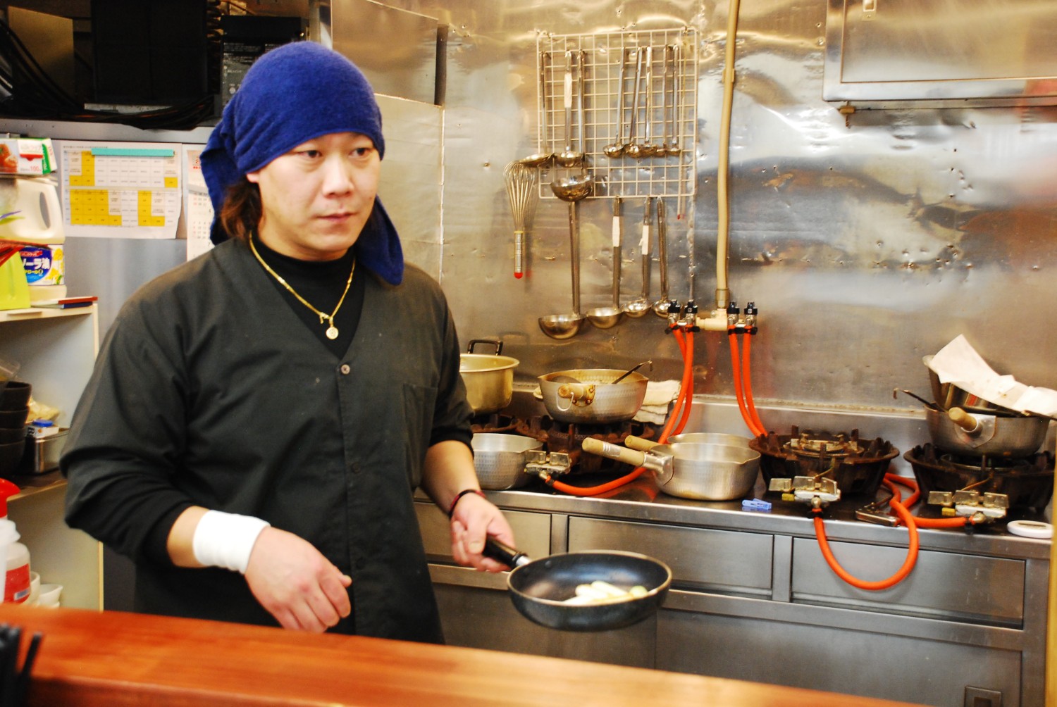 厨房の中で黒い調理服を着て青いタオルを頭に巻いた店主の阿部さんが、小さなフライパンを持って料理をしている様子の写真