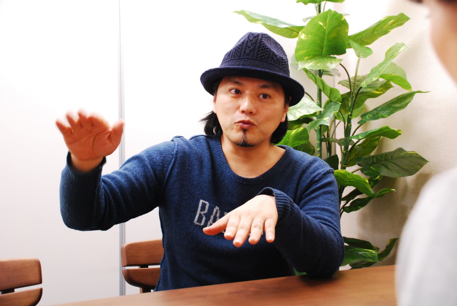 室内で青いセーターと紺の帽子を身につけた八幡さんが、手を動かしながらテーブル越しに座った人物に説明している様子の写真