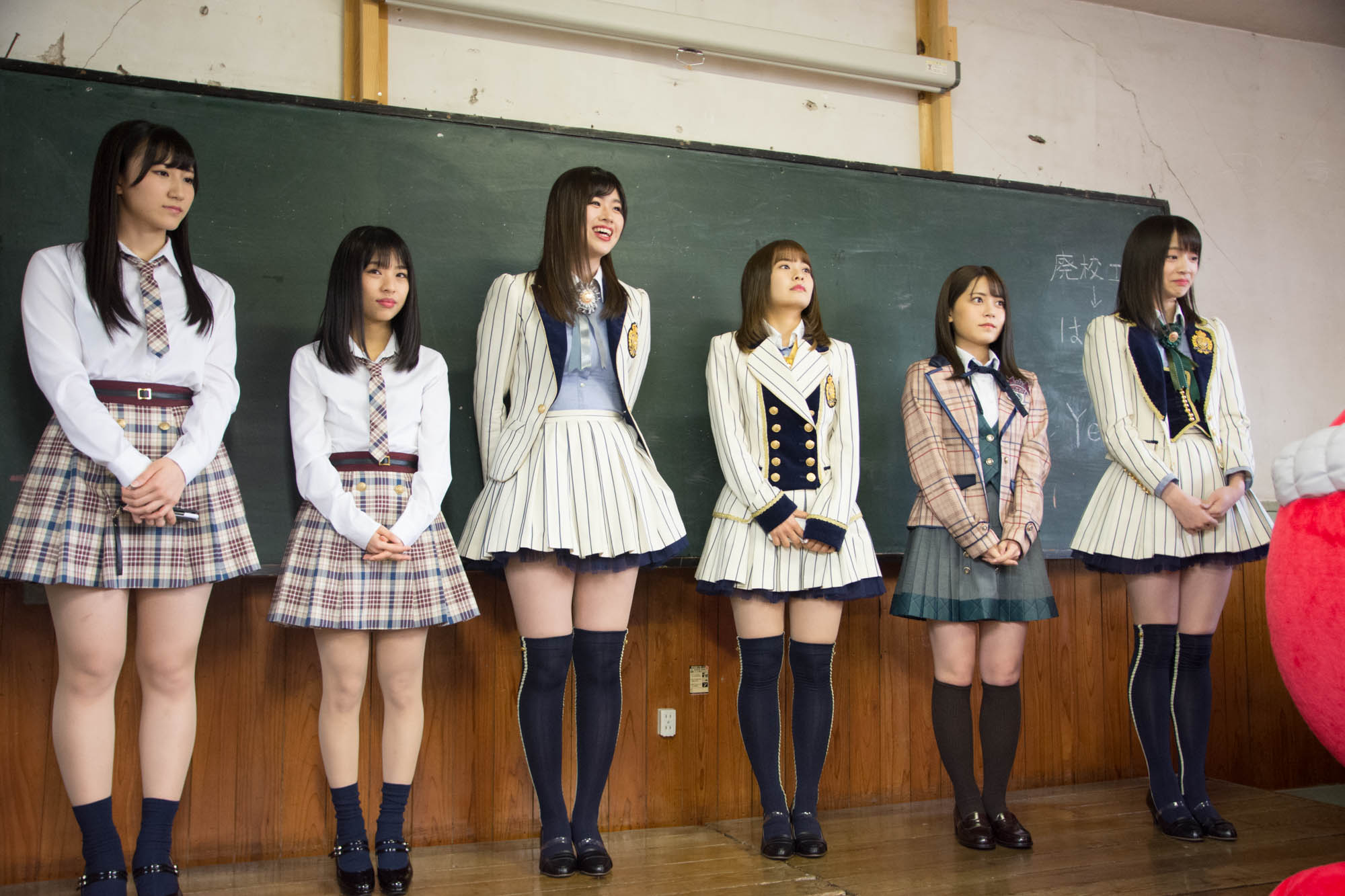 教室の黒板の前に、ストライプ柄やチェック柄の制服を着た6人のAKBグループメンバーが一列に並んで立っている写真
