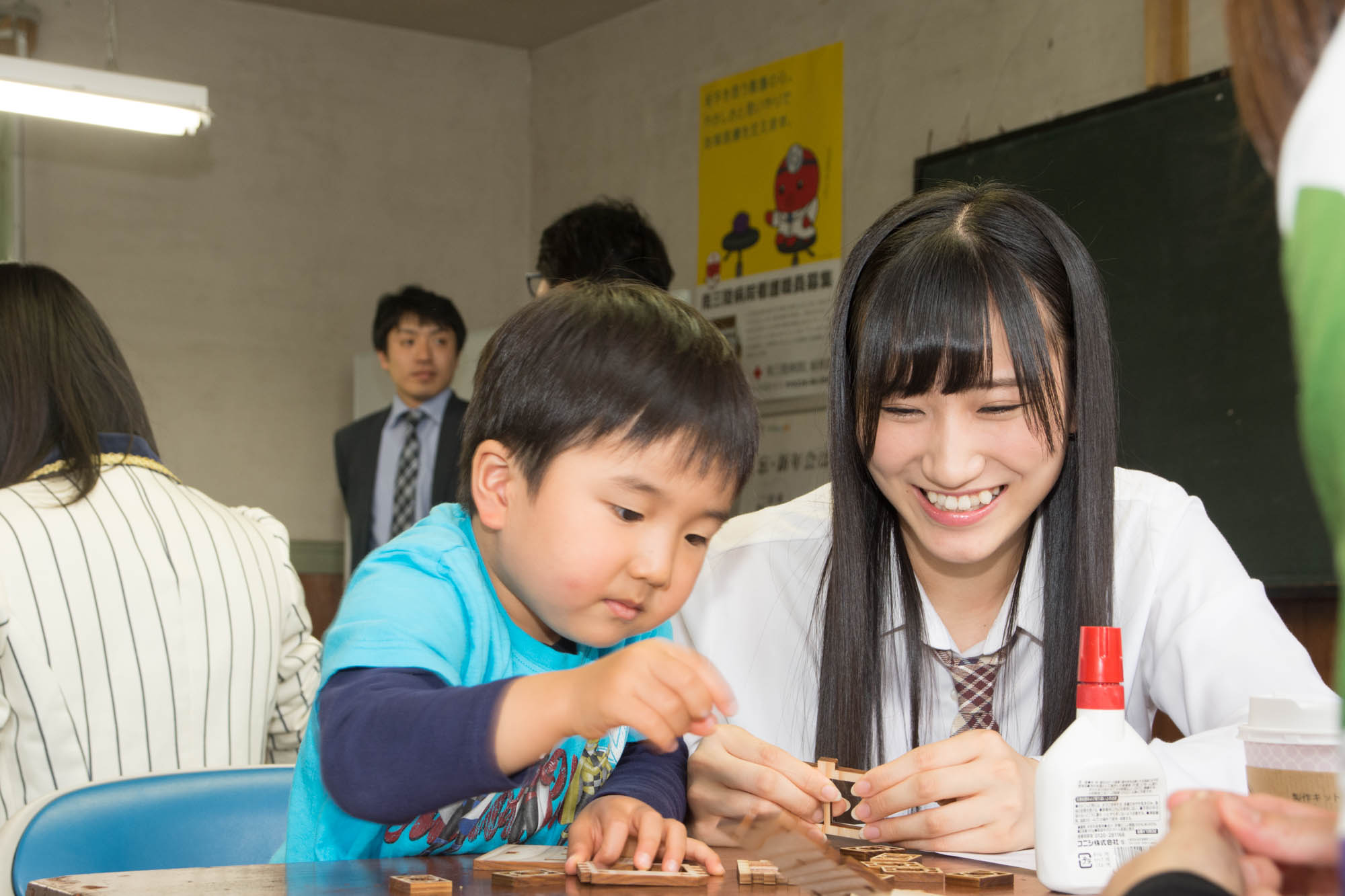 教室の机で、白いシャツとネクタイ姿の女性メンバーが小さな男の子と一緒に楽しそうに木のパーツを組み立てている写真