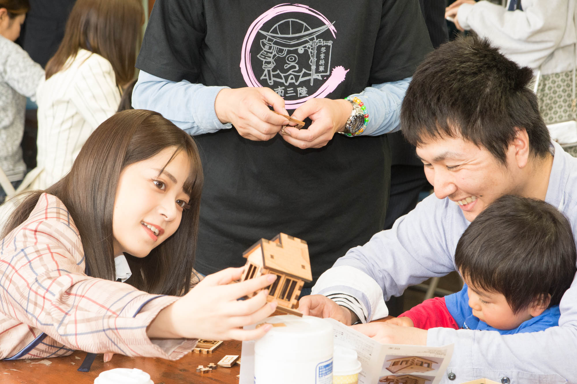 チェック柄の制服を着た女性メンバーが、男性とその膝の上にいる小さな男の子に完成した家の木製模型を見せている写真