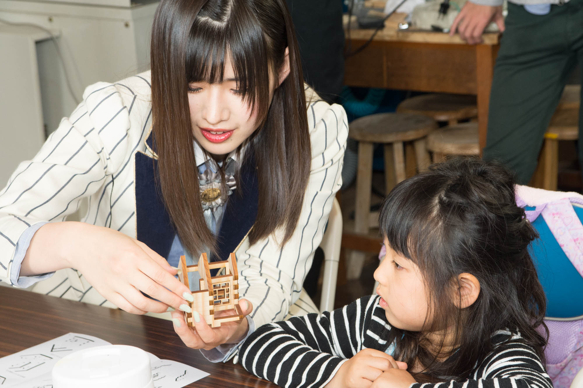 ストライプ柄の制服を着た女性メンバーが小さな女の子と一緒に木製模型を作っている写真