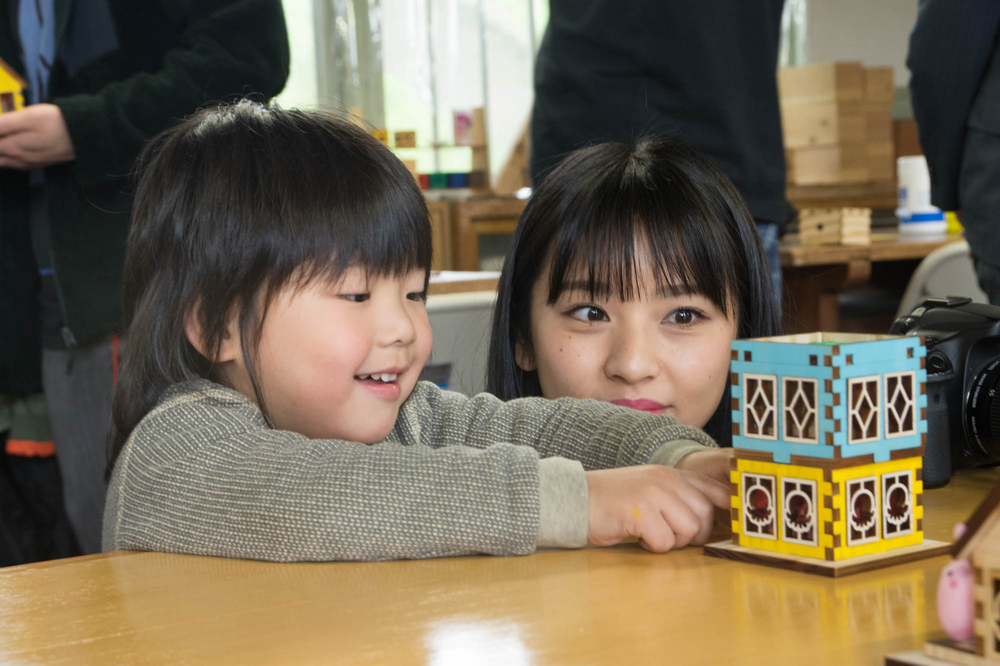 幼い子どもがテーブルの上に置かれたカラフルな木製の家の模型を2段に重ねて笑顔を見せており、横に座った女性メンバーが子どもの様子を優しく見守っている写真