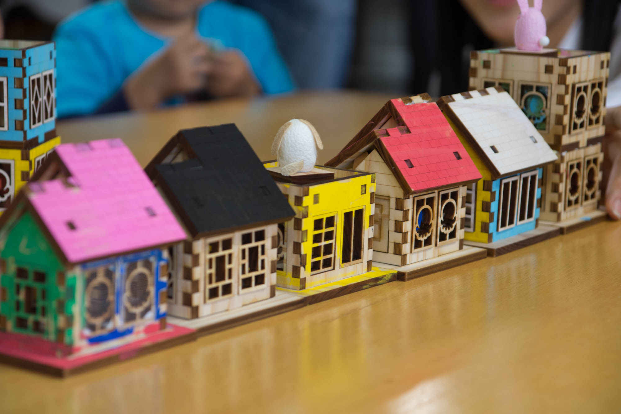 カラフルなピンク、黄色、黒、白などの屋根にまゆ人形が置かれた木製の家の模型がテーブルの上に並べられた様子をアップで撮影した写真