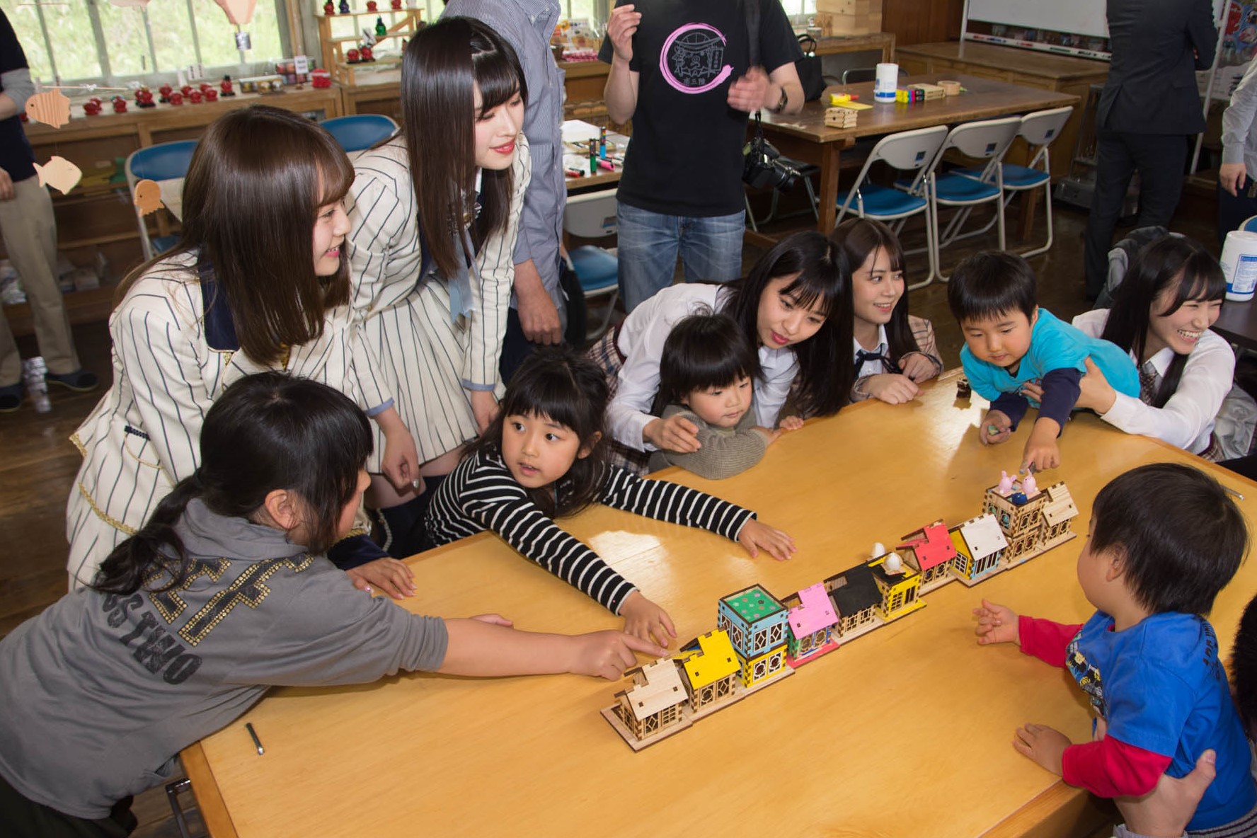 教室に世知された長テーブルを囲んで複数の子どもと2人の女性メンバーが、一列に並ばせた完成したカラフルな木製の家の模型の前で交流している写真