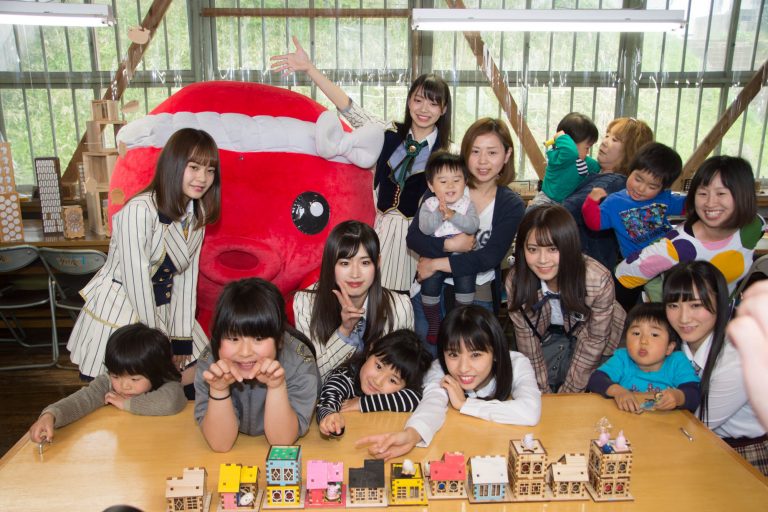 ねじり鉢巻きをしたオクトパス君とAKBのメンバーが、子どもたちと一緒に小さな木の家の工作を前にしてカメラに笑顔を向けている写真