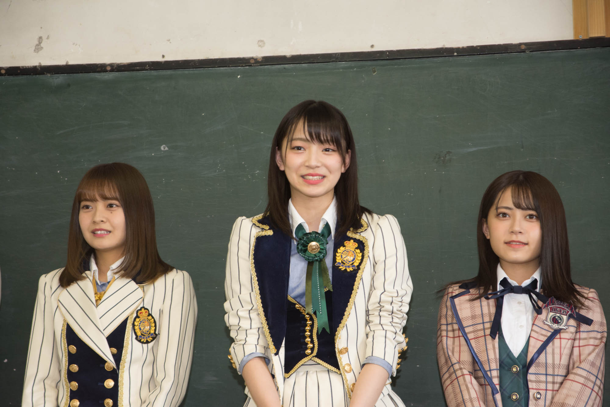 ストライプ柄やチェック柄の制服を着た3人の女性メンバーが黒板の前に並んで立ち、中央の女性が笑顔で話しをしている写真