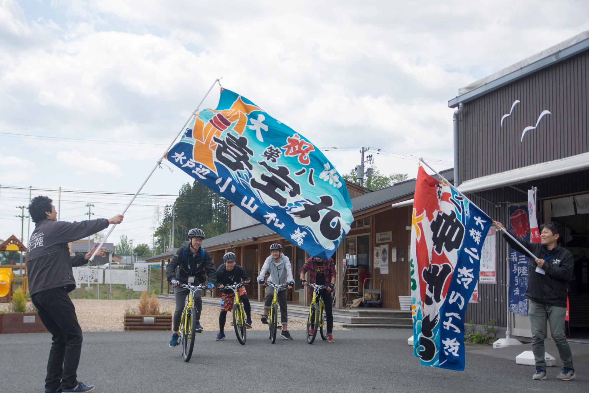 茶色の外壁の南三陸ポータルセンターの建物の前で、ヘルメットを被った4人の男性が黄色い自転車にまたがってスタートしようとしており、その手前で2人の男性スタッフが大漁旗をスタートゲートのようにして持っている写真