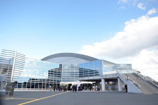 雲が浮かぶ青空の下、建物の前には数人の人々が集まり、右側に屋外階段がある大きな全面ガラス張りの建物があるベイサイドアリーナの建物外観の写真
