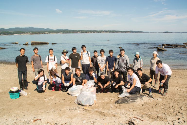 青空の下、穏やかな海が広がる砂浜で、若者を中心とした人々が笑顔で並び、中央には拾い集めた大きなゴミ袋が積まれている様子の写真