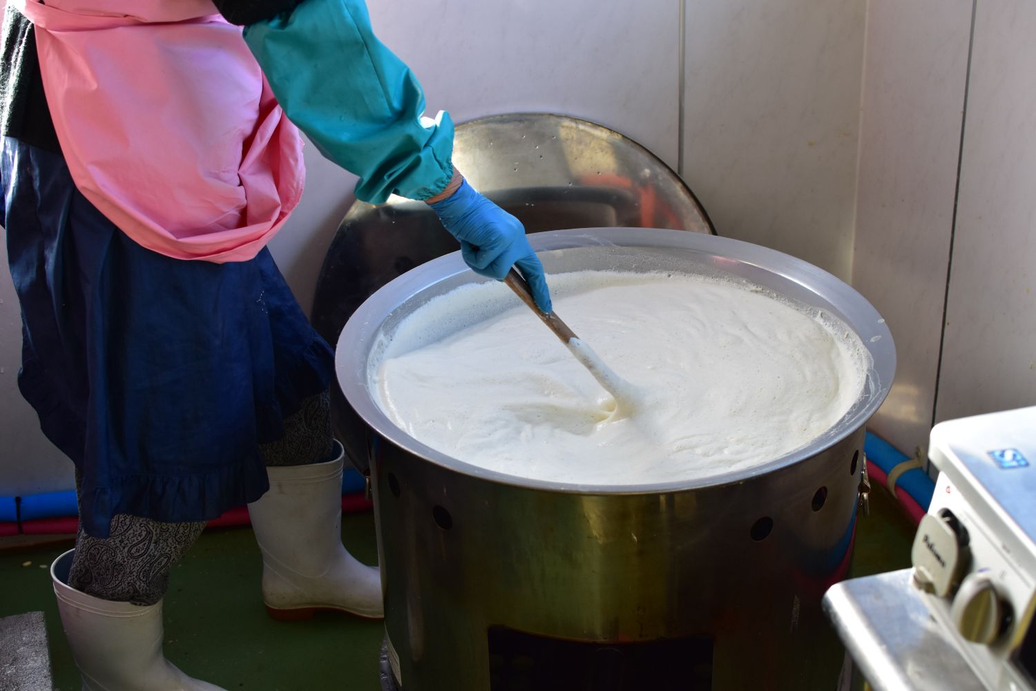 腕カバーと手袋をつけた女性が、豆を煮ている大きな鍋をかき混ぜている写真