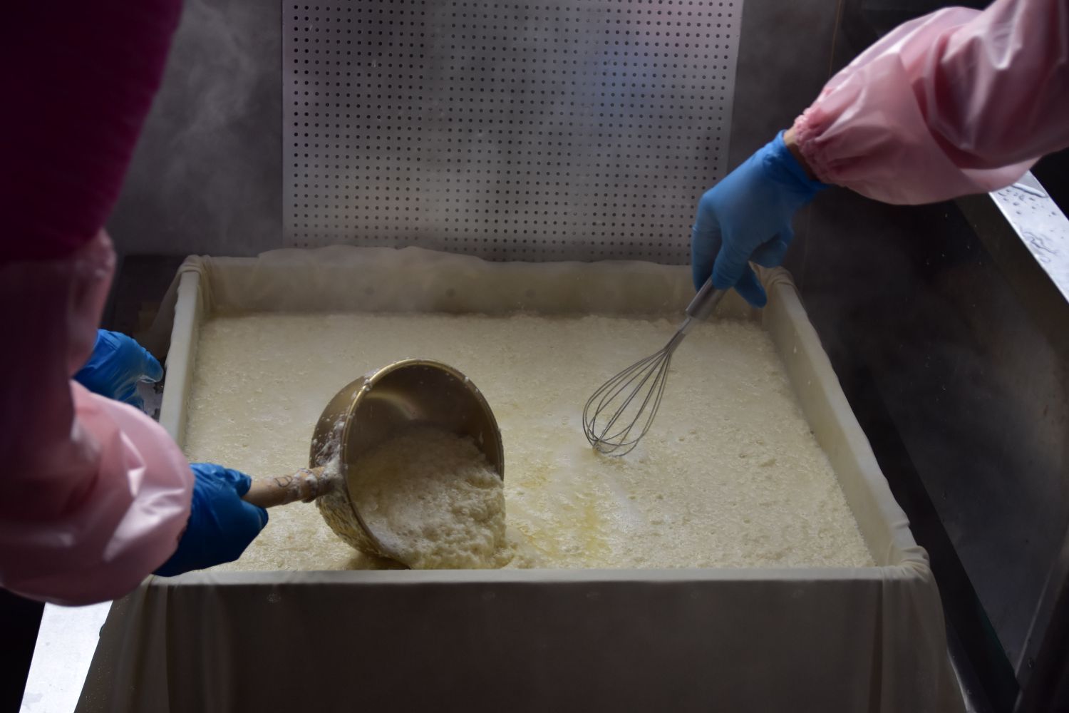 布を敷いた長方形の型に流し込んだ豆乳とにがりを、2人の女性が泡立て器や小さな鍋を使ってかき混ぜている様子の写真