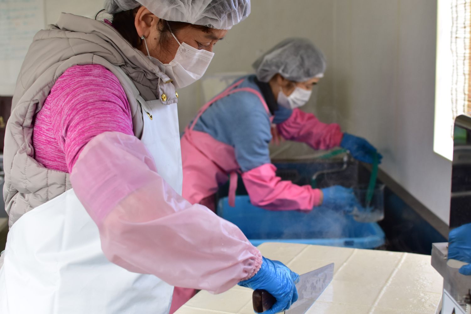 不織布キャップを被りエプロンや腕カバーをつけた女性が蛇口で水をくみ、手前の女性は完成した豆腐に包丁を使ってカットしている様子の写真