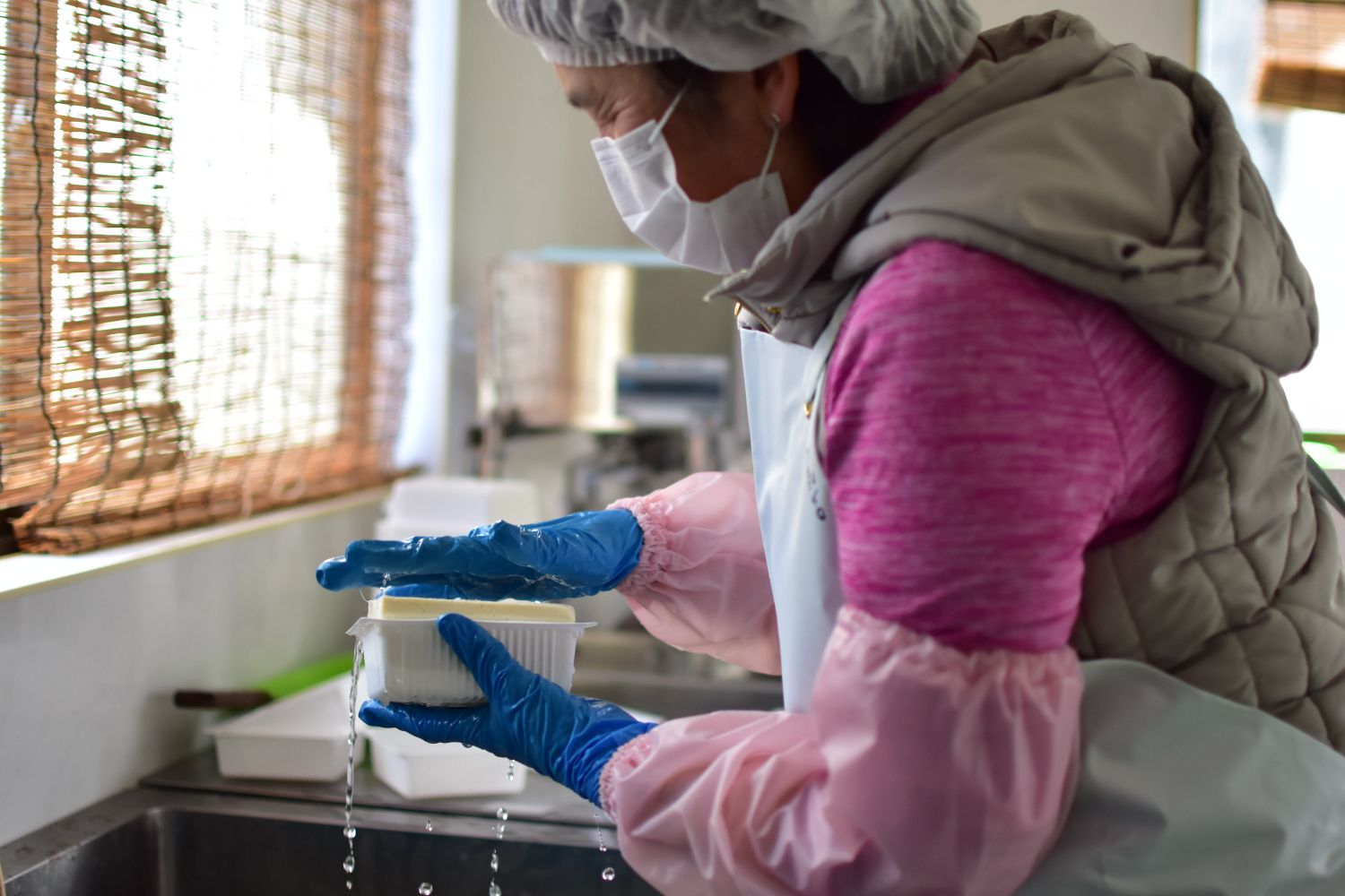 腕カバーを着け、手袋をはめた女性の作業者が、白い容器に完成した豆腐を入れている様子の写真