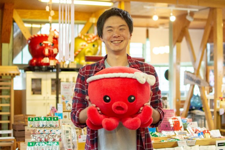木製の梁がある明るい店内に並んだ商品の前で、ねじり鉢巻きをしたタコのぬいぐるみを笑顔で両手で持つ、赤のチェック柄のシャツを着た大森さんをアップで撮影した写真