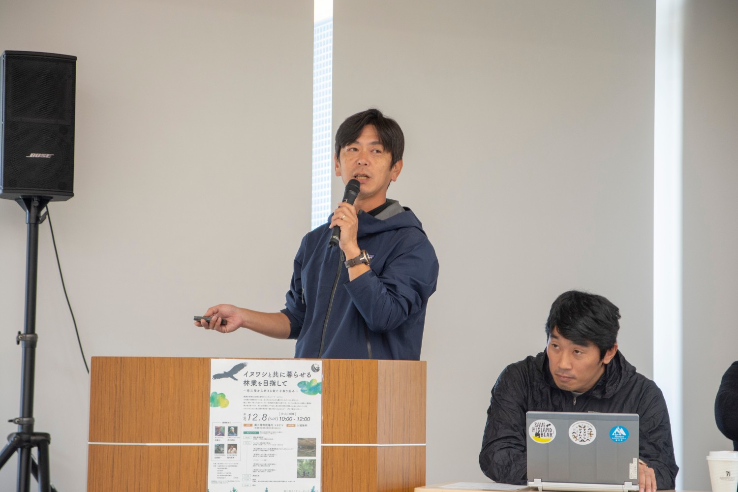 演壇に立ってマイクを持ちながら話している人物と、その隣でノートパソコンを操作している人物が写っている写真