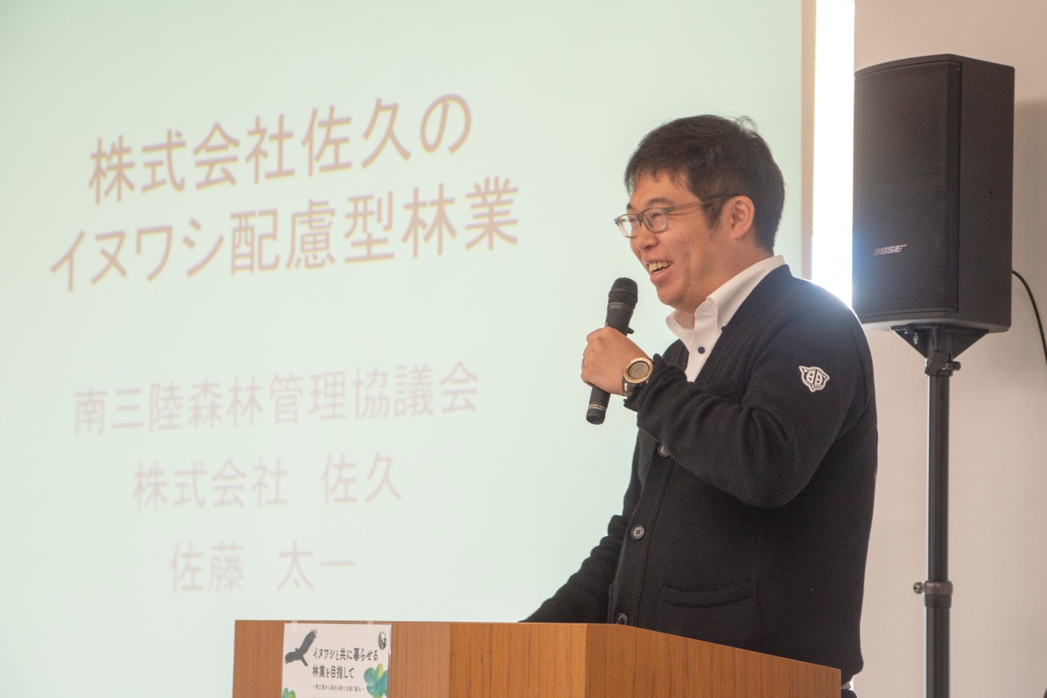 スクリーンに「株式会社佐久のイヌワシ配慮型林業 南三陸森林管理協議会 株式会社佐久 佐藤太一」と書かれた文字が中央に大きく表示され、その横の演題でマイクを持った佐藤さんが話をしている写真