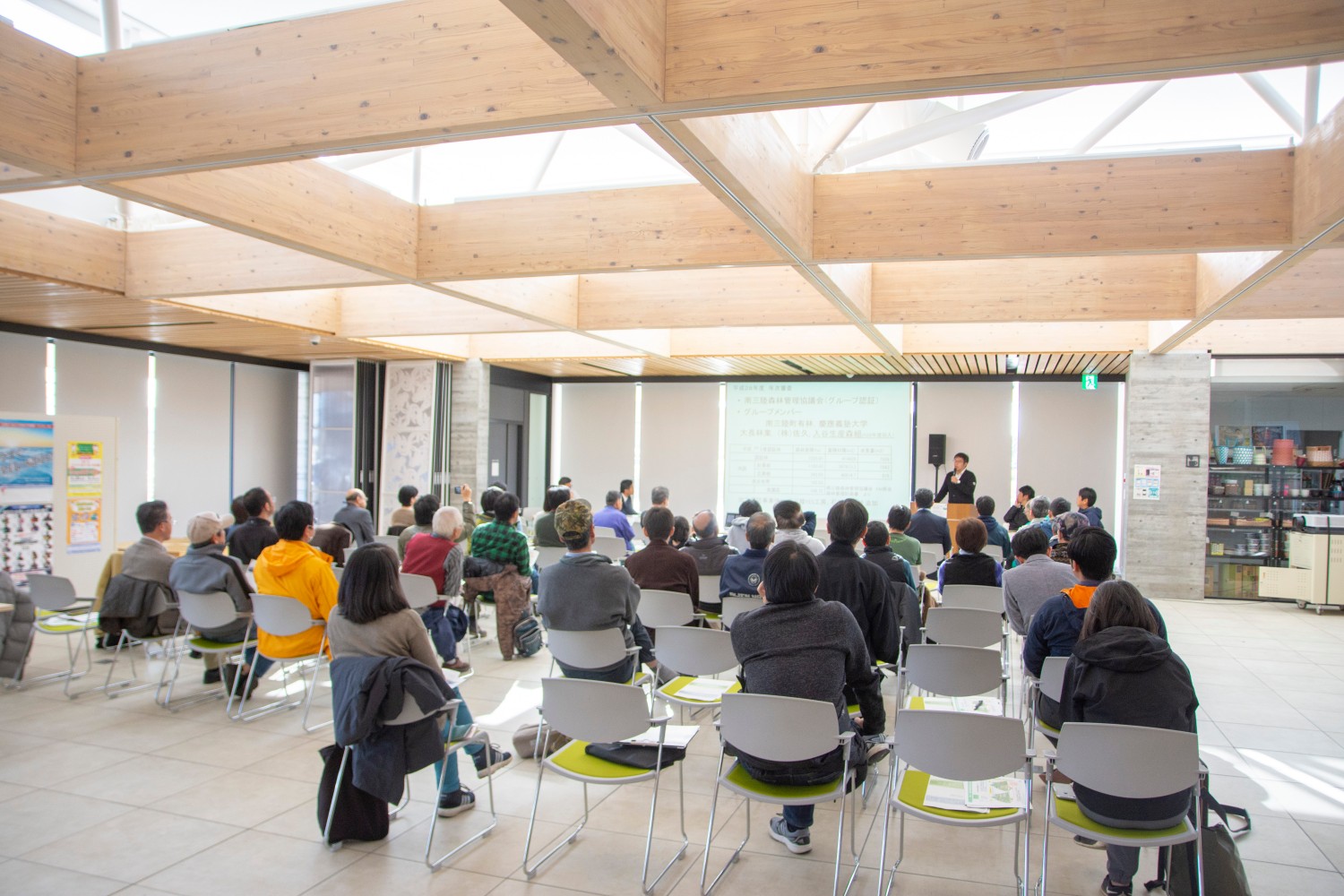 明るい室内で多数の人々が椅子に座り、前方のスクリーンを見ながら講義を受けている様子が写っている写真