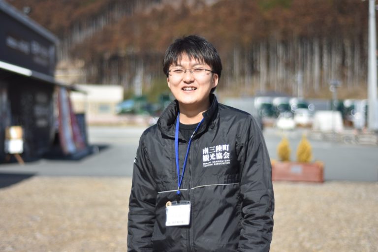 右側に沢山の木々が立つ林と駐車したトラックと建物を背景に、左胸に「南三陸町観光協会」と書かれた黒いジャンパーを着た阿部さんをアップで撮影した写真