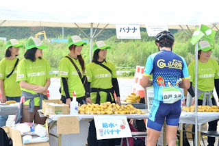緑色のTシャツと帽子を着た女性たちがテントの下でバナナを並べており、自転車のユニフォーム姿の参加者が「給水・バナナ」と書かれたテーブルの前に立っている補給所の写真
