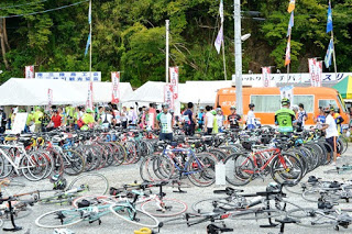 大量のロードバイクが整然と並べられた駐輪スペースに多くの参加者やスタッフが集まり、後方には白いテントやのぼり、オレンジ色の移動販売車が並ぶ写真