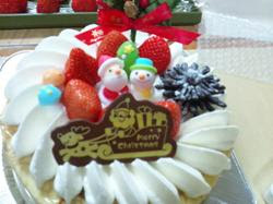 生クリームでデコレーションされた、イチゴ、サンタクロースと雪だるまの飾り、松ぼっくり、『Merry Christmas』と書かれたチョコレートプレートが載っているクリスマスケーキの写真