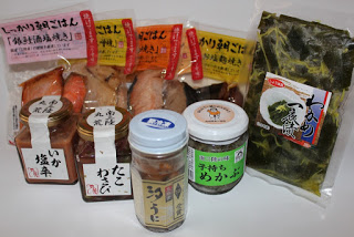 汐うに、子持ちめかぶ、たこわさび、いか塩辛の4つの瓶の後ろに真空パックに入った魚の切り身の商品4つとわかめ一夜漬のパックが並んでいる写真