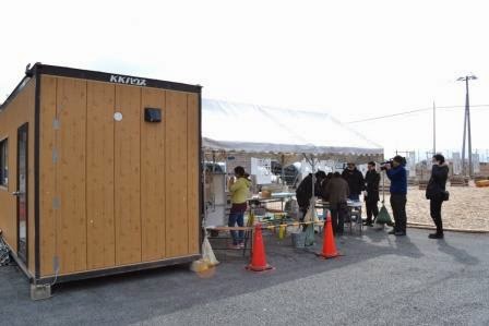 木目調のプレハブ小屋の隣に設置された白いテントに複数の人が集まって作業をしている様子の写真