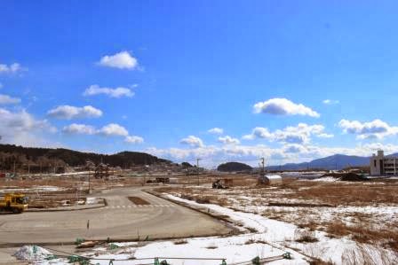 雪がまだ残る広大な更地に舗装された道路や重機が見え、遠くの山々や青空の下で復興工事が進められている様子を写した被災地の冬の風景の写真