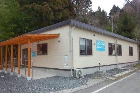 木々に囲まれた場所に建つ、クリーム色の外壁に水色の看板が設置された、木造の屋根付き通路がある建物外観の写真