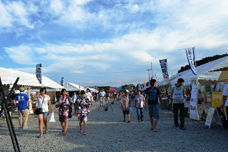 屋外の祭り会場で、多くの人がテントの間の通路を歩いており、子どもから大人まで浴衣や祭り衣装を着ている様子が見える、昼間の祭りの写真