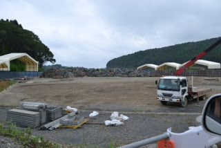 広い砂利の敷地に大きな岩や石が積まれ、白いテント状の構造物やクレーン付きトラック、資材が配置された工事現場の写真