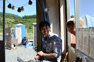 日差しの差し込む建物の入り口で若い男性が椅子に座って笑顔を見せており、横にある窓から年配の関係者が顔をのぞかせている写真