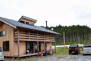 木造の二階建ての建物の前で大人と子ども数人が集まっており、背景に緑の森が広がっている写真